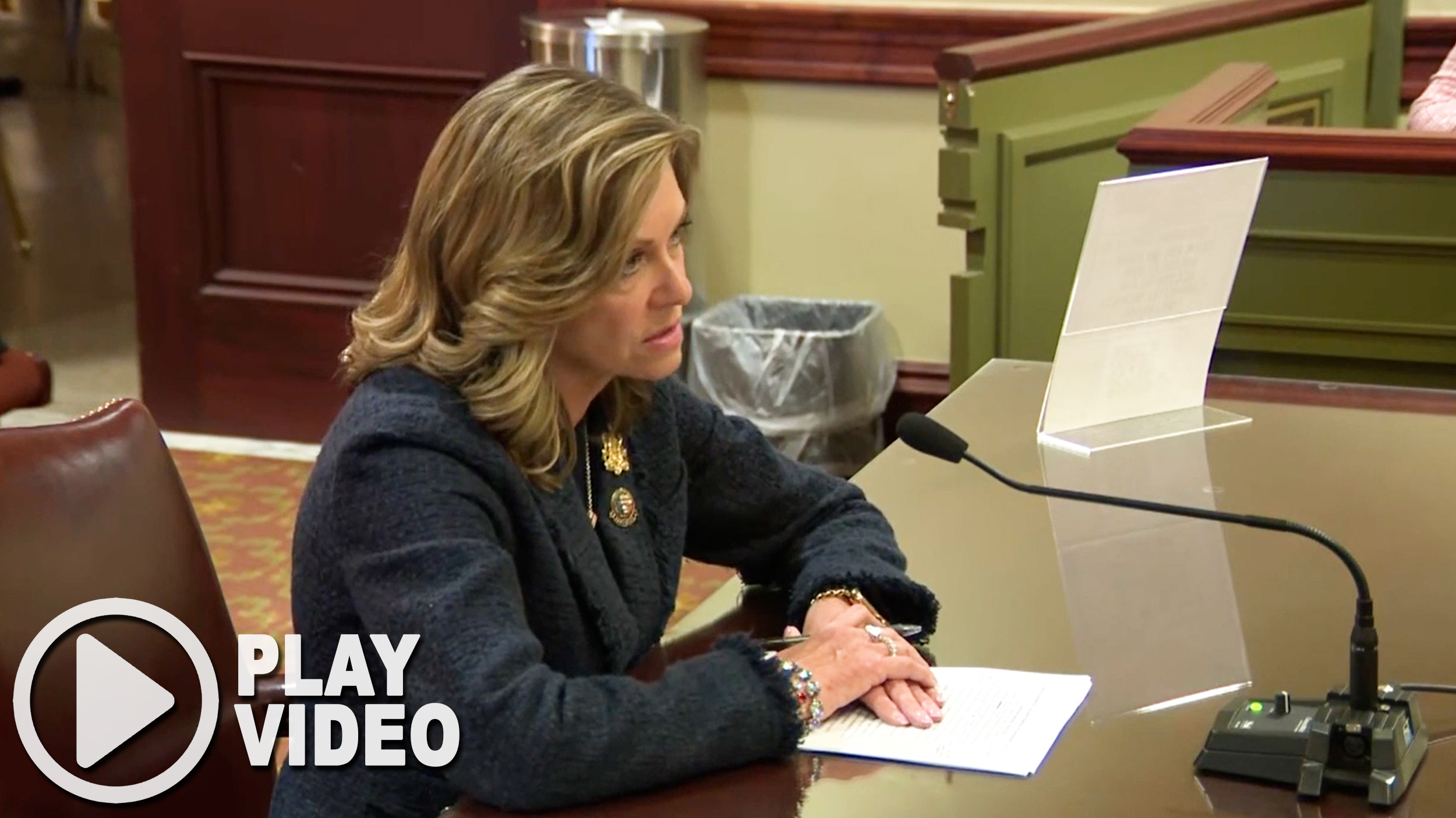 Sen. Bartolotta sits at a committee table in the Pennsylvania Capitol speaking into a microphone while presenting testimony on her Senate bill. She is seated with papers in front of her, and the image includes a “Play Video” button linking to a YouTube video of her remarks.