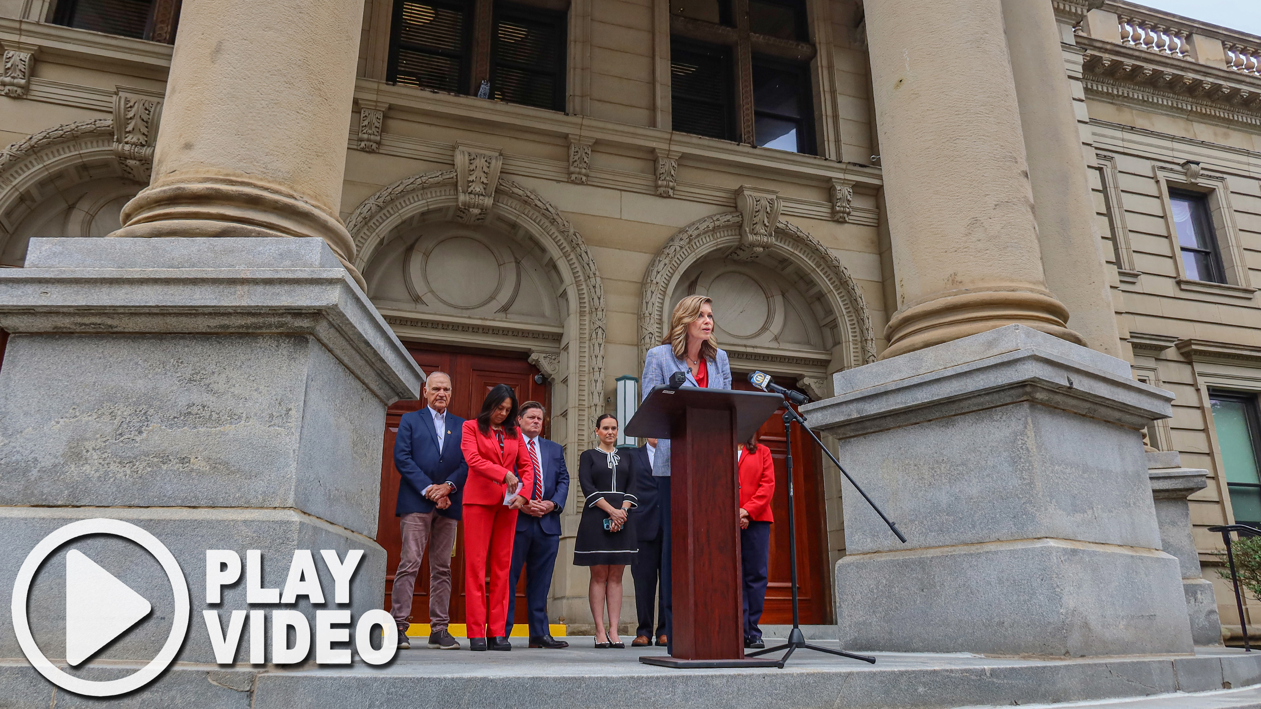 Sen. Bartolotta speaks at a podium at the top of the Washington County Courthouse front steps. A play button is in the bottom lefthand corner, indicating this image is a link to watch a video of Sen. Bartolotta’s remarks.