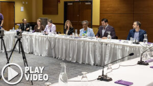 Senator Bartolotta sits with other legislators, educators, and health care leaders at a long table setup for a discussion. A play button graphic overlay is in the bottom lefthand corner of the image.