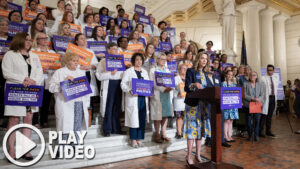 Sen. Bartolotta speaks at a podium in front of a large group of nurses and advocates holding signs as they rally in support of Senate Bill 25 and House Bill 739. A graphic overlay of a play button is in the bottom lefthand corner of the image.