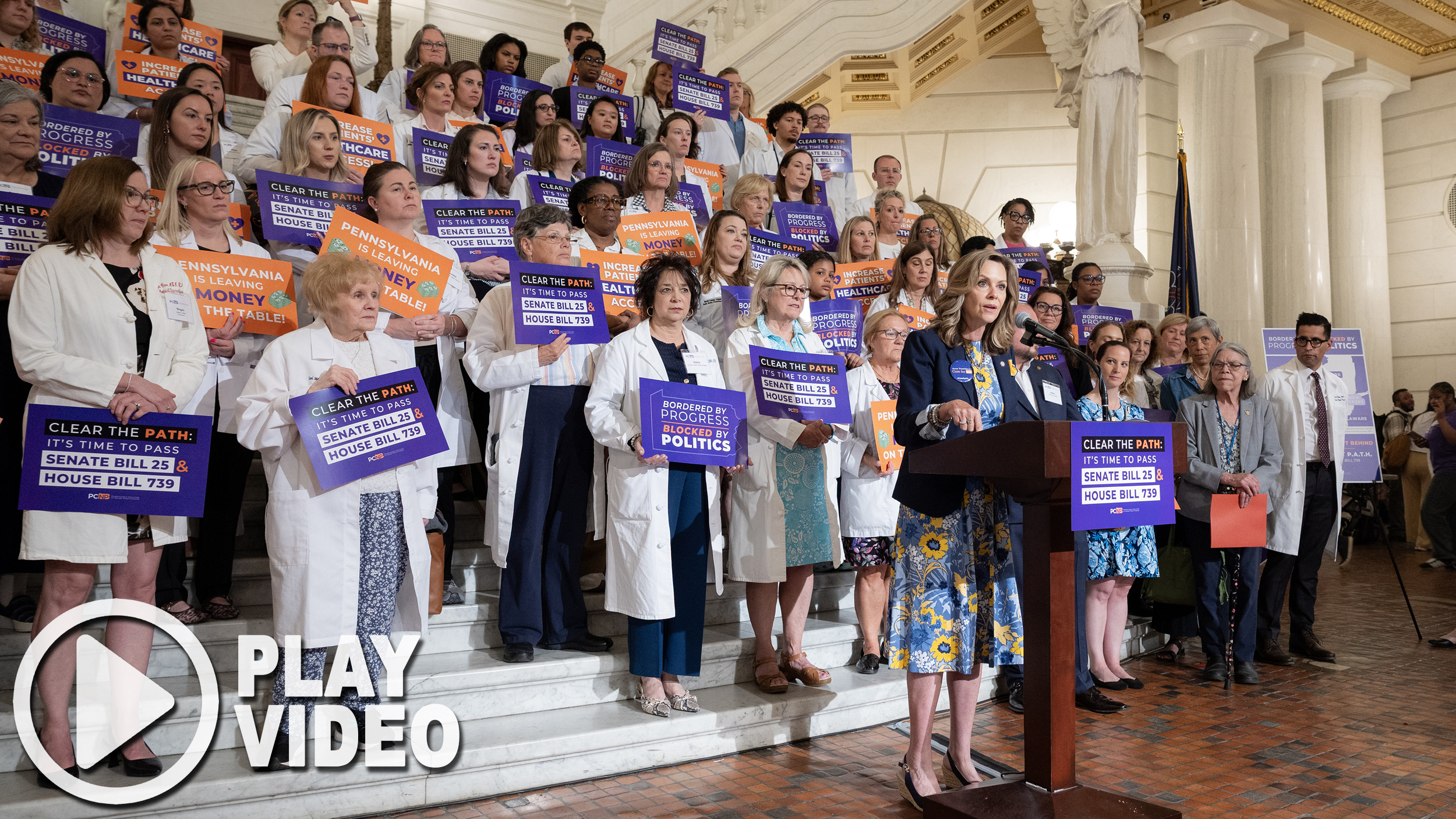 Sen. Bartolotta speaks at a podium in front of a large group of nurses and advocates holding signs as they rally in support of Senate Bill 25 and House Bill 739. A graphic overlay of a play button is in the bottom lefthand corner of the image.