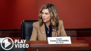 Sen. Bartolotta looks to her right while seated during a committee hearing. A graphic overlay of a play button is in the bottom lefthand corner of the image.