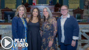 Sen. Bartolotta poses with Marlee Solomon, her mother, Daneen, and Coach Sheila Mitchell-Rodi. A graphic overlay of a play button is in the bottom lefthand corner of the image.