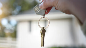 Hand dangling house keys with white home in background.